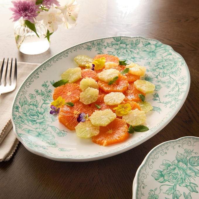 A floral-patterned plate with slices of orange and grapefruit, garnished with edible flowers and mint leaves, sits on a wooden table next to a fork, napkin, and a small vase of fresh flowers.