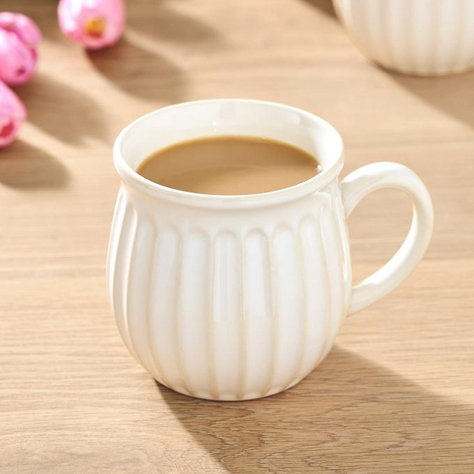A white ceramic mug filled with coffee sits on a wooden table next to pink and yellow tulips. Other similar mugs are in the background.