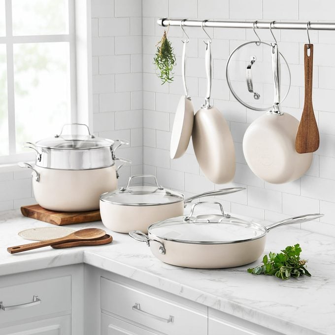 A set of cream-colored pots and pans with glass lids arranged on a white marble kitchen counter, with some cookware and lids hanging from a metal rack above. Fresh herbs and wooden utensils are nearby.