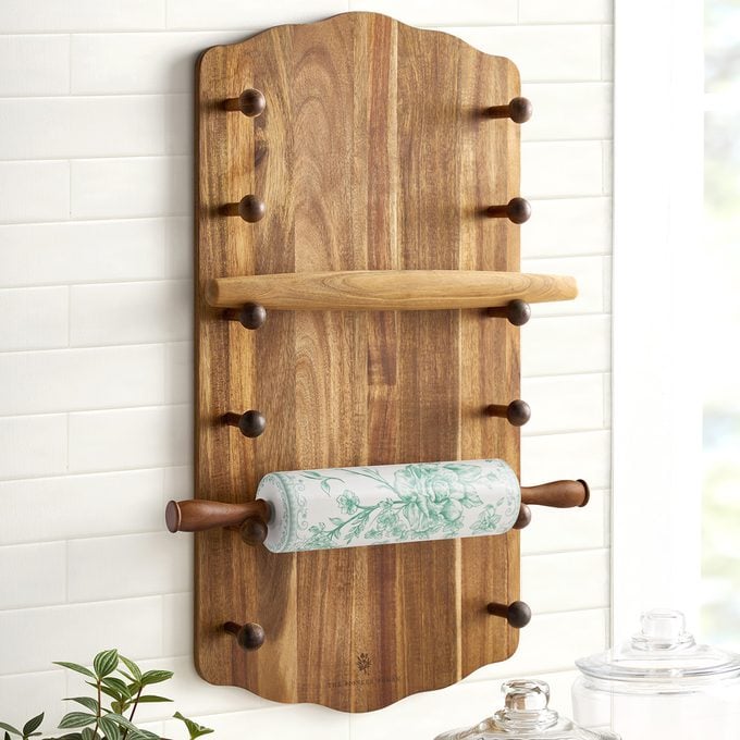 A wooden wall-mounted organizer with pegs holds a rolling pin and decorative paper, set against a white tiled wall. Nearby, glass jars and green plant leaves add to the organized, inviting space.